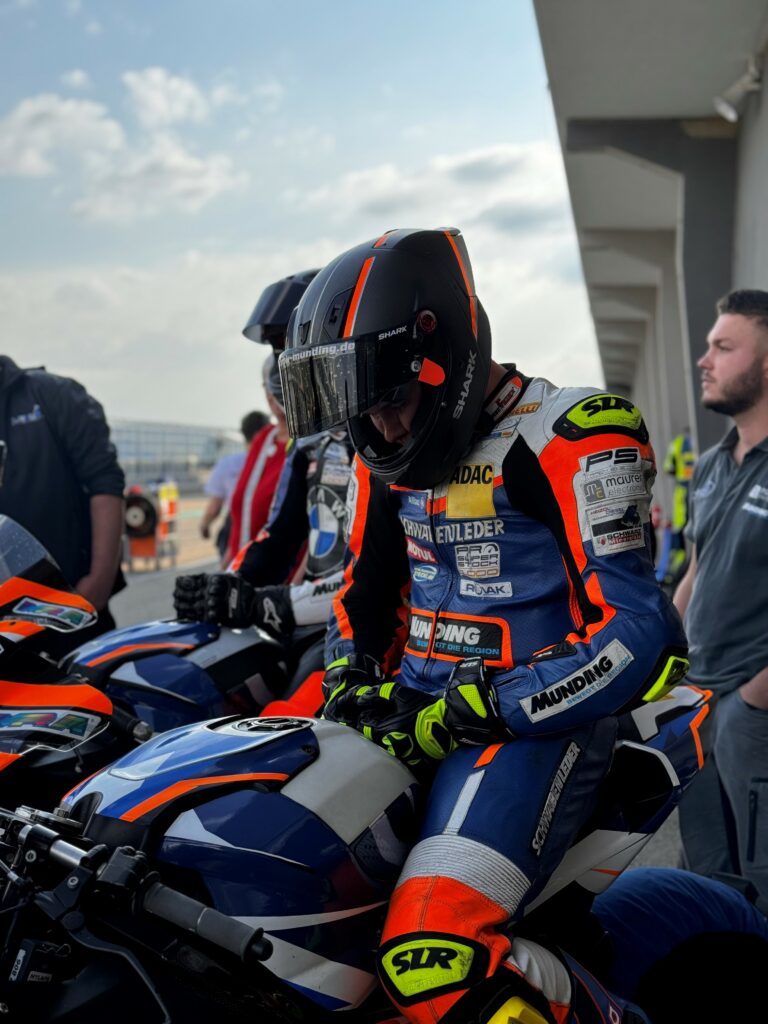 Im Vordergrund sitzt eine Person auf einem Motorrad welche Orange Blau gekleidet ist. Daneben sitzt eine Person ebenfalls auf dem Motorrad. Links und Rechts von den Rennfahrern stehen 2 Personen die Grau gekleidet sind.
