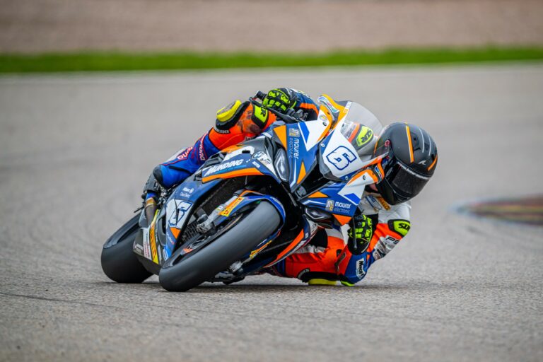 Blau, Orange farbige BMW S1000RR f&auml;hrt auf dem Sachsenring in Schr&auml;glage durch eine Kurve, diese f&auml;hrt eine Person welche in den gleichen Farben eine Lederkombi tr&auml;gt.