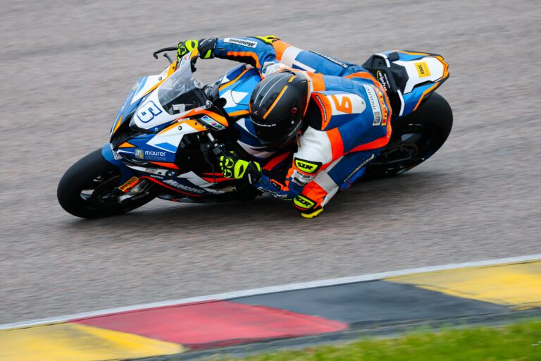 Blau, Orange farbige BMW S1000RR f&auml;hrt auf dem Sachsenring in Schr&auml;glage durch eine Kurve, diese f&auml;hrt eine Person welche in den gleichen Farben eine Lederkombi tr&auml;gt.